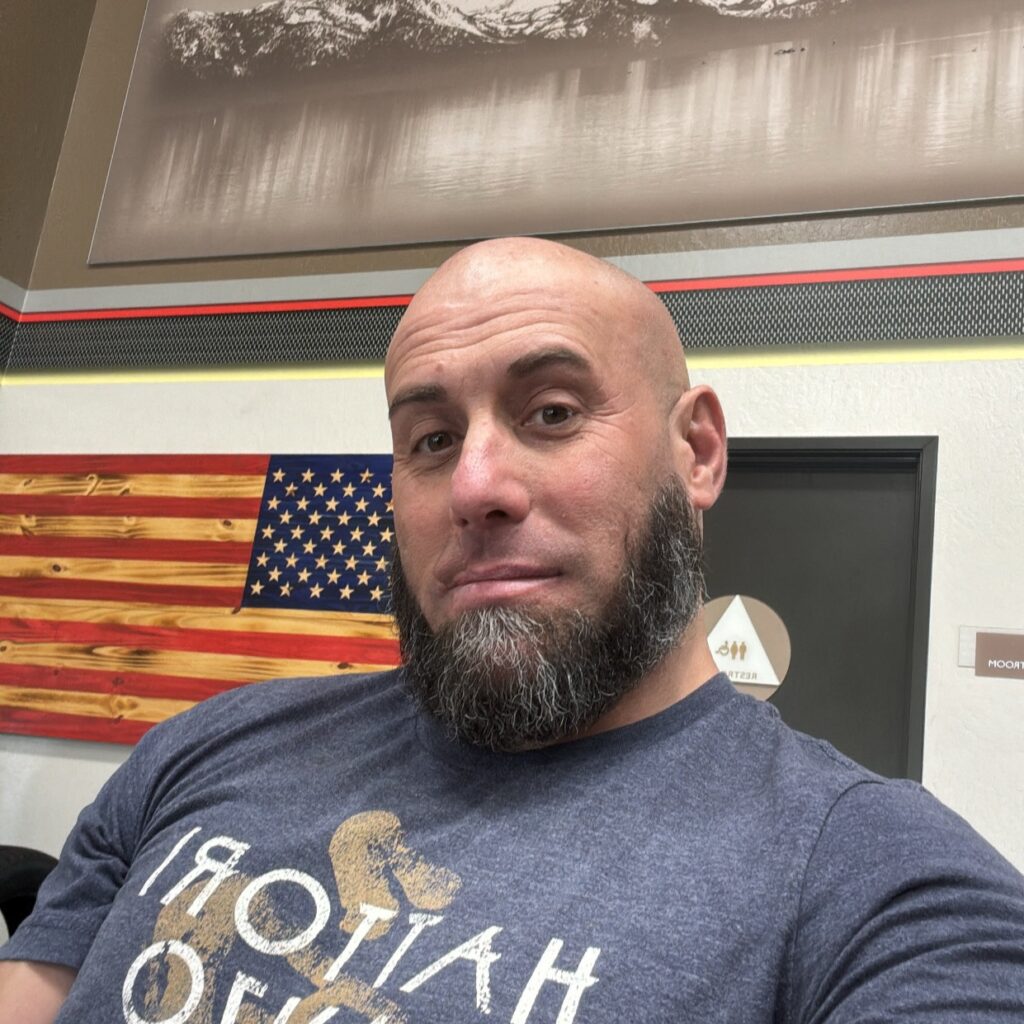 Close-up of a bald man with a gray beard taking a selfie in front of a wooden American flag wall art.