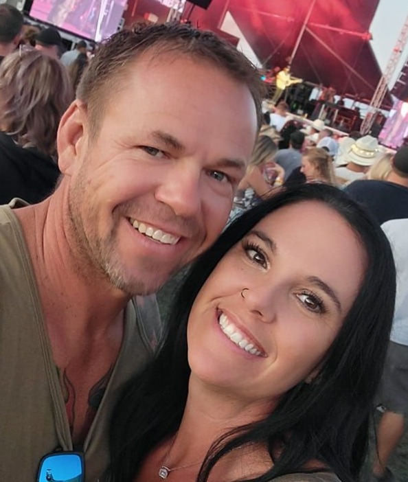 Smiling couple taking a selfie at an outdoor concert, with a crowd and red stage backdrop in the background (man on left, woman on right).